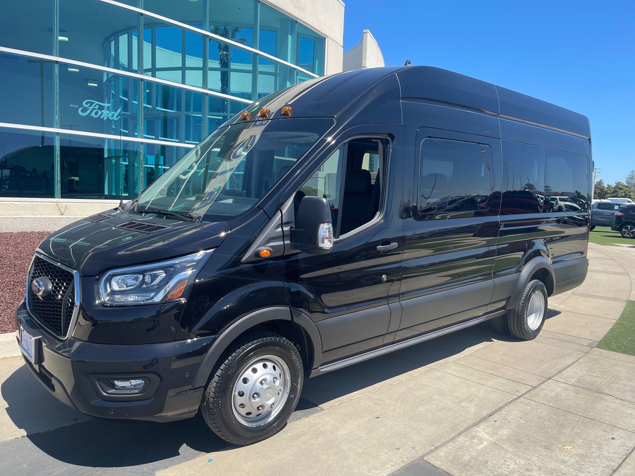 Photograph of a Ford Transit shuttle. It is a black vehicle parked on a sunny day.